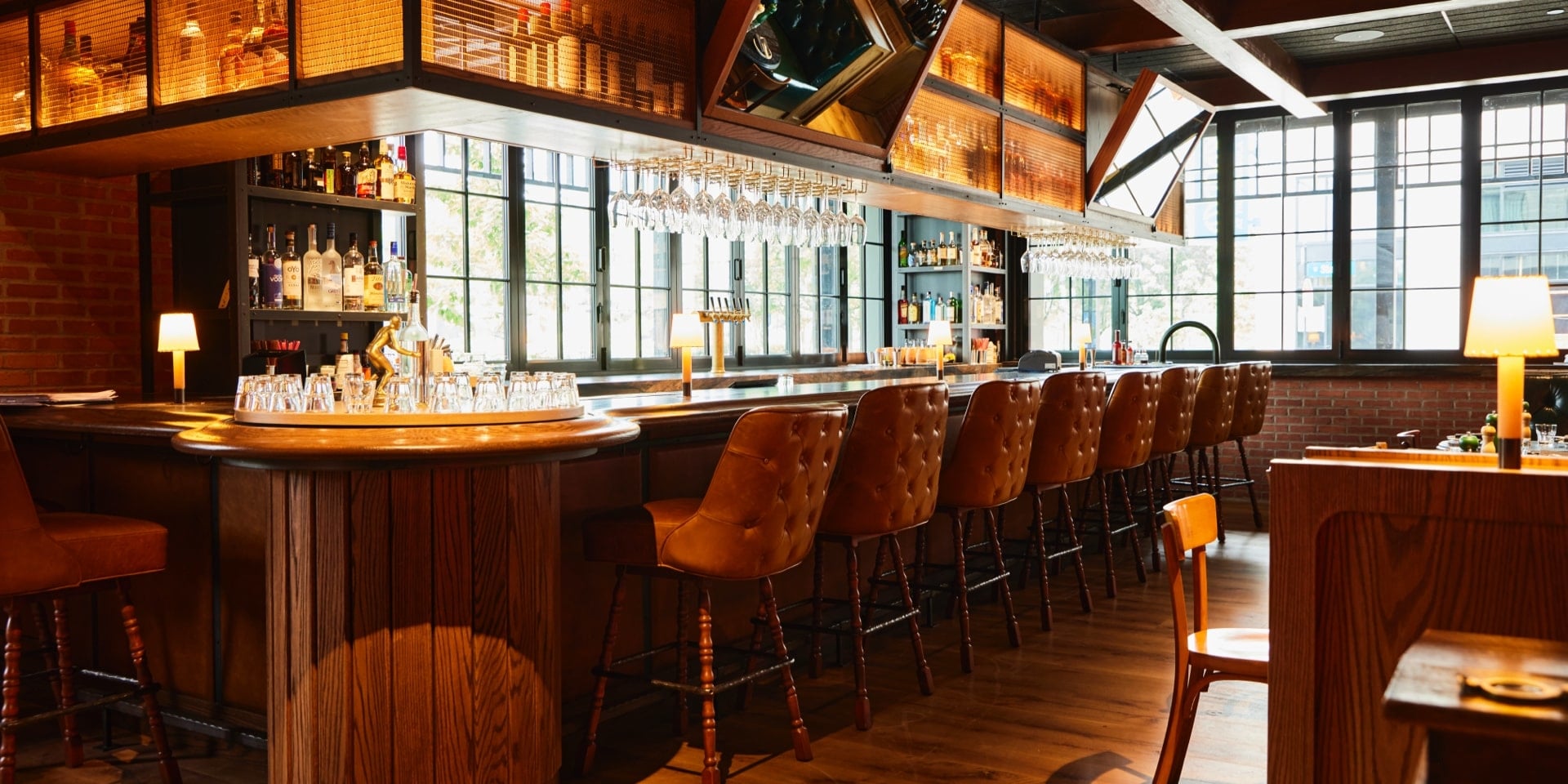 Wood-paneled bar with tufted leather stools, hanging wine glasses, and exposed brick at Little West Tavern, Columbus