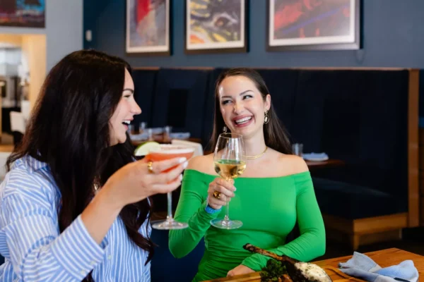 Two women laughing and toasting with a pink cocktail and white wine at a restaurant table at Margaritaville Hotel Nashville